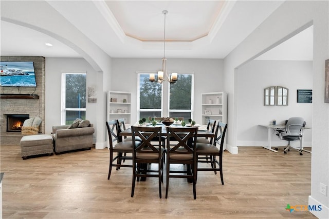 dining room featuring a stone fireplace, ornamental molding, a tray ceiling, a notable chandelier, and light hardwood / wood-style floors