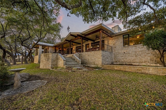 back of property at dusk featuring a yard, stairway, stone siding, and a ceiling fan