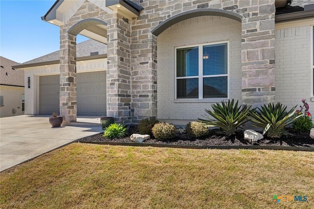 doorway to property with concrete driveway, stone siding, a garage, a yard, and a shingled roof