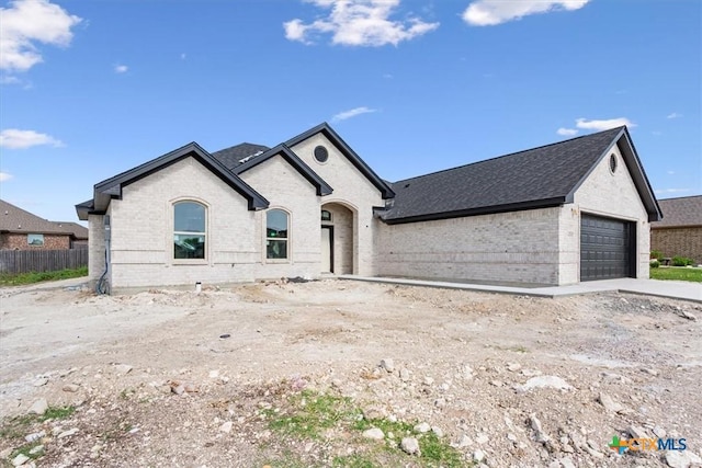 french provincial home with brick siding, an attached garage, roof with shingles, and fence