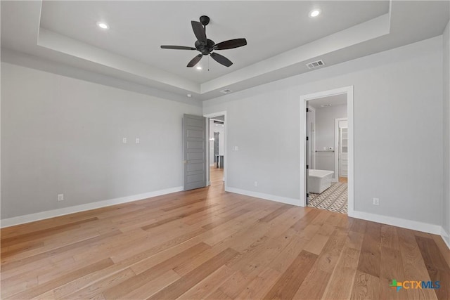 unfurnished bedroom featuring ensuite bathroom, ceiling fan, a tray ceiling, and light hardwood / wood-style flooring