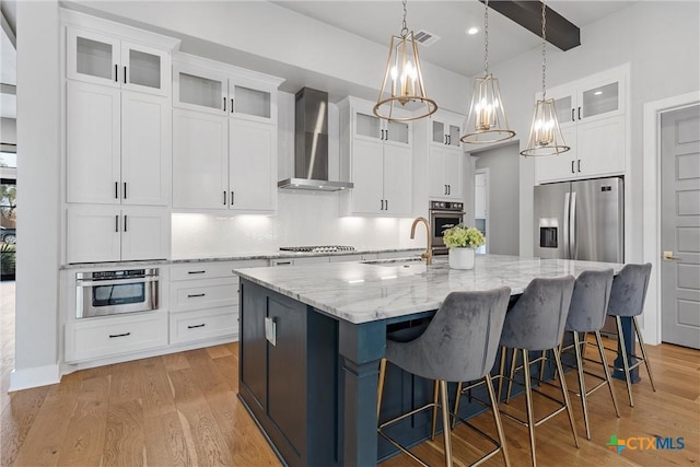 kitchen featuring an island with sink, stainless steel appliances, wall chimney range hood, white cabinets, and sink