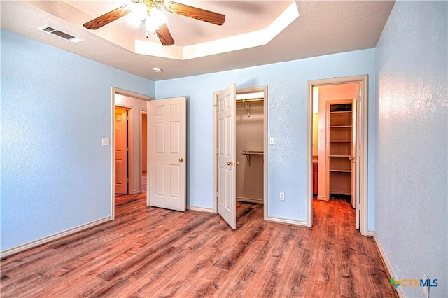 unfurnished bedroom with a spacious closet, a tray ceiling, wood finished floors, and visible vents