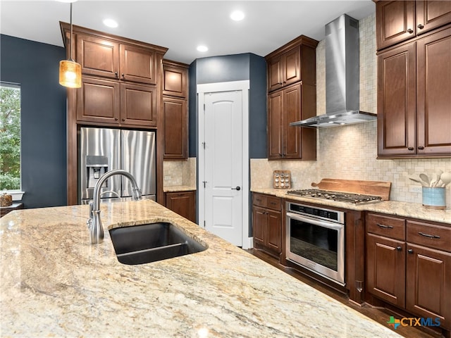 kitchen featuring sink, hanging light fixtures, stainless steel appliances, light stone countertops, and wall chimney range hood