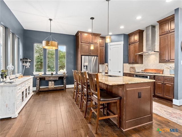 kitchen with sink, stainless steel fridge with ice dispenser, light stone countertops, a kitchen island with sink, and wall chimney range hood