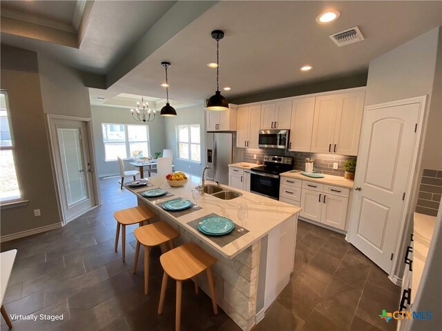 kitchen with stainless steel appliances, a center island with sink, sink, white cabinetry, and decorative light fixtures