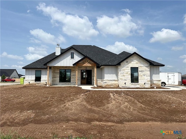 modern farmhouse style home with board and batten siding, roof with shingles, a chimney, and stone siding
