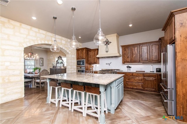 kitchen featuring light parquet floors, hanging light fixtures, an island with sink, appliances with stainless steel finishes, and light stone counters