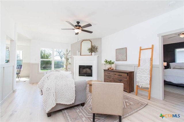 bedroom with light wood-type flooring, a glass covered fireplace, a ceiling fan, and wainscoting