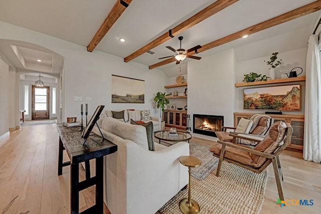 living room featuring beam ceiling, a warm lit fireplace, light wood-type flooring, arched walkways, and ceiling fan