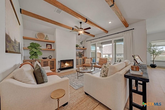 living area featuring a fireplace with flush hearth, visible vents, light wood-style flooring, beamed ceiling, and ceiling fan