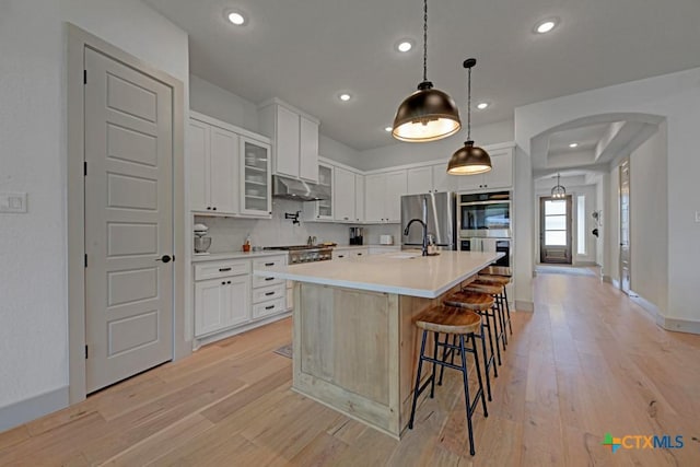 kitchen with light countertops, stainless steel appliances, under cabinet range hood, arched walkways, and light wood-style flooring