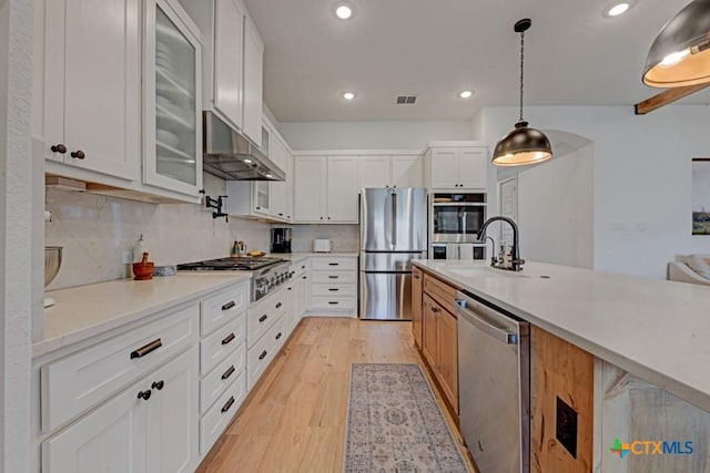 kitchen featuring appliances with stainless steel finishes, under cabinet range hood, glass insert cabinets, a sink, and light wood-style floors