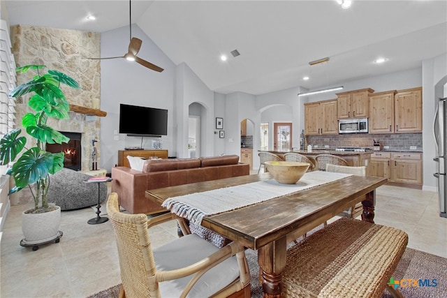 dining area featuring arched walkways, high vaulted ceiling, a stone fireplace, light tile patterned floors, and a ceiling fan