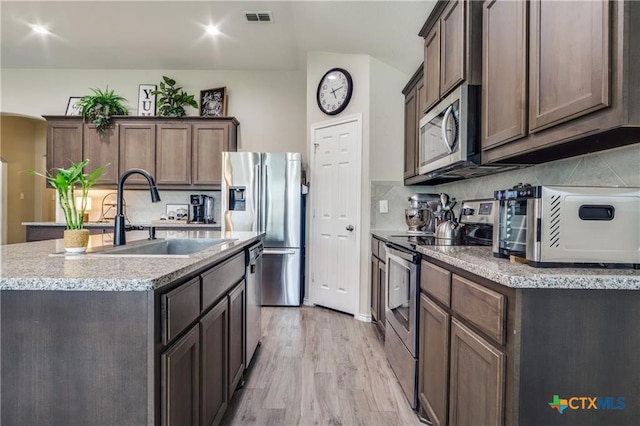 kitchen featuring stainless steel appliances, sink, light stone counters, a kitchen island with sink, and light hardwood / wood-style flooring