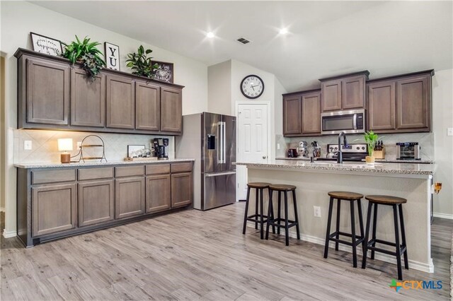 kitchen with stainless steel appliances, light stone countertops, a kitchen island with sink, and light wood-type flooring