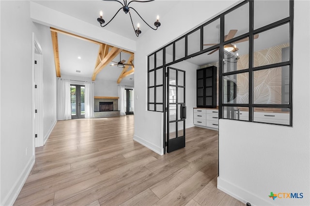 unfurnished living room with french doors, ceiling fan with notable chandelier, a premium fireplace, beam ceiling, and light hardwood / wood-style floors