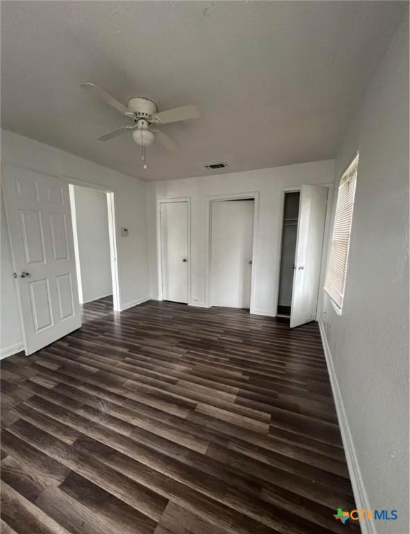 unfurnished bedroom featuring ceiling fan, visible vents, baseboards, two closets, and dark wood-style floors