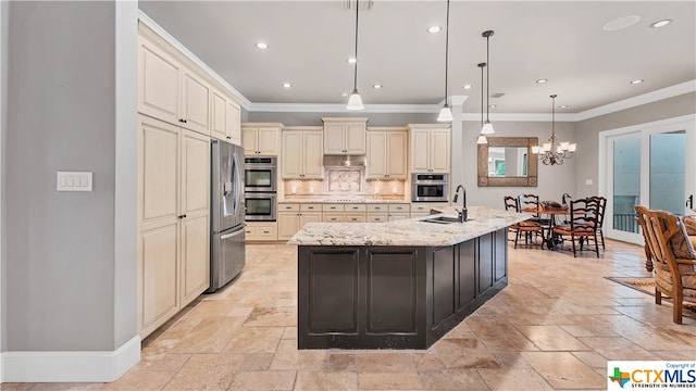 kitchen with appliances with stainless steel finishes, light stone countertops, a kitchen island with sink, cream cabinetry, and pendant lighting