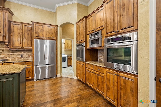 kitchen featuring appliances with stainless steel finishes, decorative backsplash, dark wood-type flooring, light stone countertops, and ornamental molding