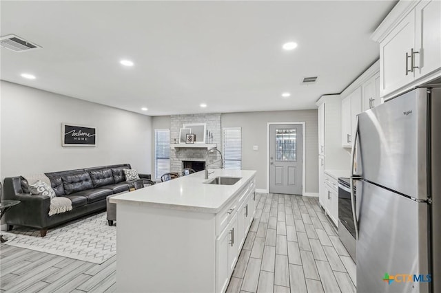 kitchen with stainless steel fridge, a brick fireplace, sink, a center island with sink, and white cabinetry