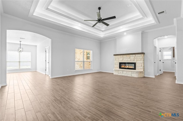 unfurnished living room featuring a raised ceiling, ceiling fan, a stone fireplace, and crown molding