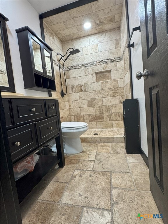 bathroom with toilet, stone tile flooring, tiled shower, and vanity
