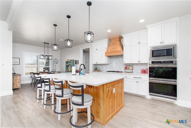 kitchen featuring premium range hood, a kitchen island with sink, white cabinets, decorative light fixtures, and stainless steel appliances