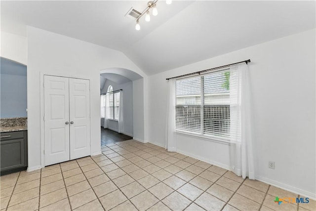 empty room featuring arched walkways, lofted ceiling, healthy amount of natural light, light tile patterned flooring, and baseboards