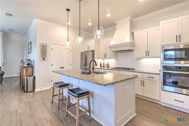 kitchen with white cabinetry, custom range hood, stainless steel appliances, light stone countertops, and a kitchen island with sink