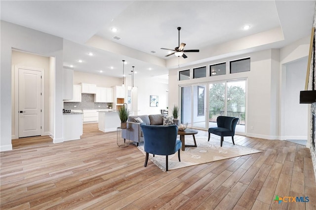 living room with a raised ceiling, ceiling fan, a stone fireplace, and light hardwood / wood-style floors