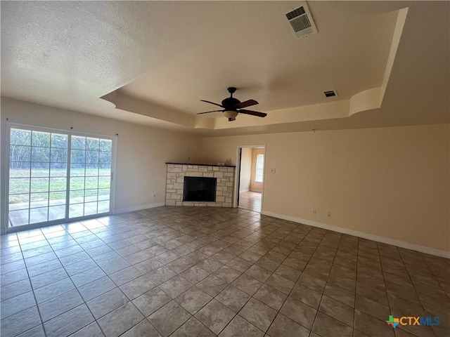 unfurnished living room with ceiling fan, a tray ceiling, a stone fireplace, and a wealth of natural light