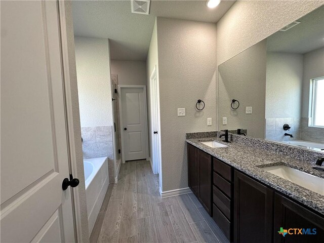 bathroom with a textured wall, a garden tub, wood finished floors, baseboards, and double vanity
