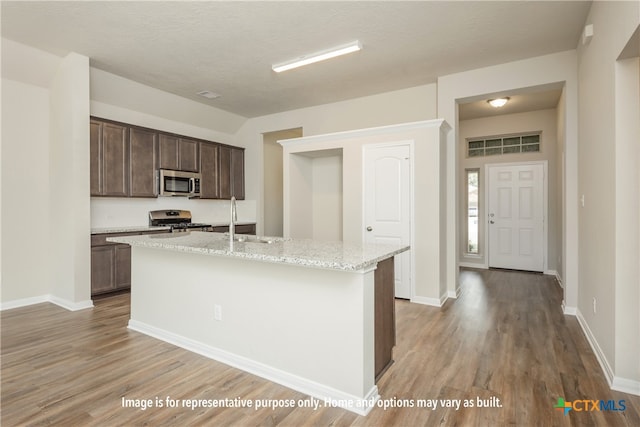 kitchen with dark brown cabinetry, appliances with stainless steel finishes, light stone countertops, an island with sink, and light hardwood / wood-style flooring