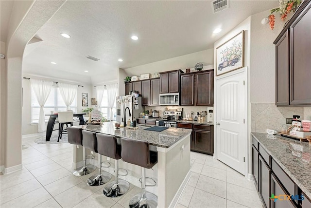 kitchen featuring appliances with stainless steel finishes, light stone counters, dark brown cabinets, a kitchen island with sink, and a breakfast bar area