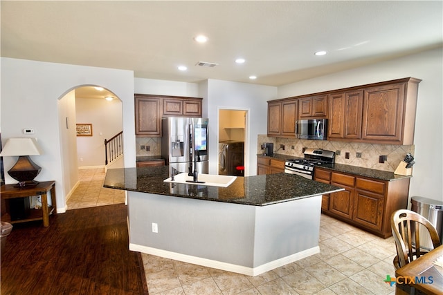 kitchen with appliances with stainless steel finishes, light hardwood / wood-style floors, dark stone countertops, and an island with sink