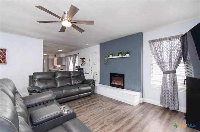 living area with a glass covered fireplace, ceiling fan, baseboards, and wood finished floors