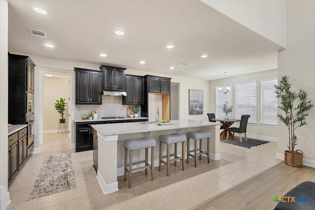 kitchen featuring tasteful backsplash, light stone counters, a kitchen bar, a center island with sink, and light wood-type flooring