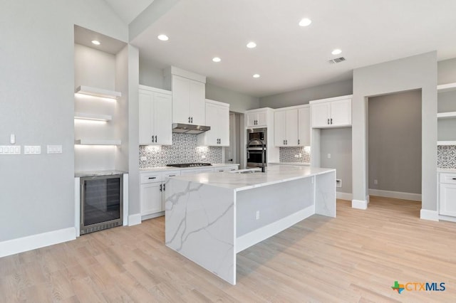 kitchen with wine cooler, light stone counters, light hardwood / wood-style floors, white cabinetry, and an island with sink