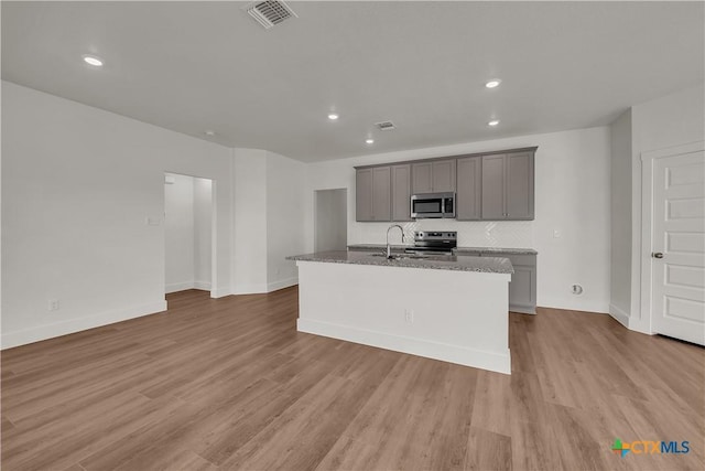 kitchen with gray cabinets, an island with sink, light stone counters, stainless steel appliances, and light wood-type flooring