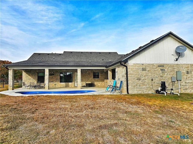 rear view of property featuring stone siding, a lawn, roof with shingles, and a patio area