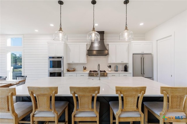 kitchen with custom exhaust hood, stainless steel appliances, a large island with sink, white cabinetry, and hanging light fixtures