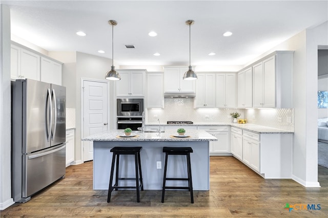 kitchen featuring appliances with stainless steel finishes, white cabinets, a center island with sink, and decorative light fixtures