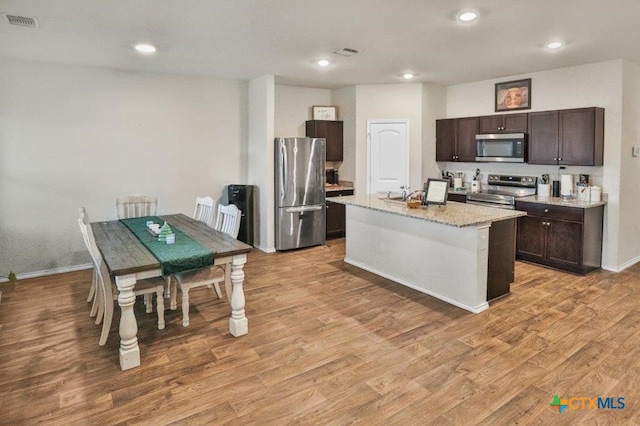 kitchen with appliances with stainless steel finishes, dark brown cabinetry, a kitchen island with sink, sink, and light hardwood / wood-style flooring