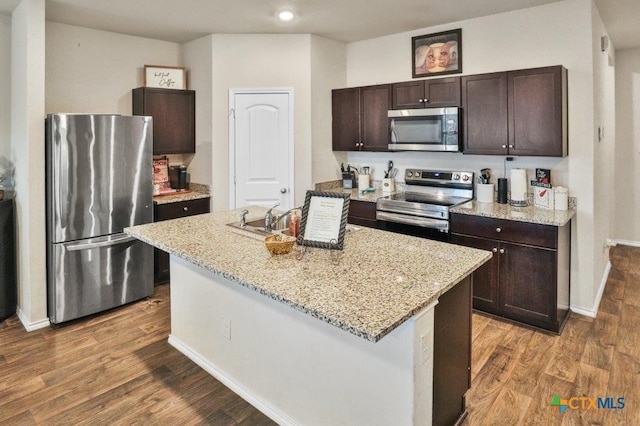 kitchen featuring dark brown cabinetry, light stone countertops, an island with sink, and appliances with stainless steel finishes