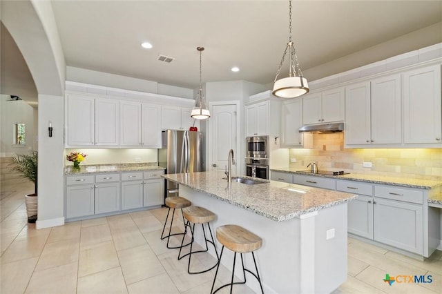 kitchen featuring pendant lighting, sink, stainless steel appliances, light stone counters, and a center island with sink