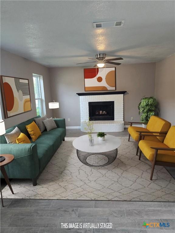 living room featuring a ceiling fan, a textured ceiling, visible vents, a brick fireplace, and wood finished floors