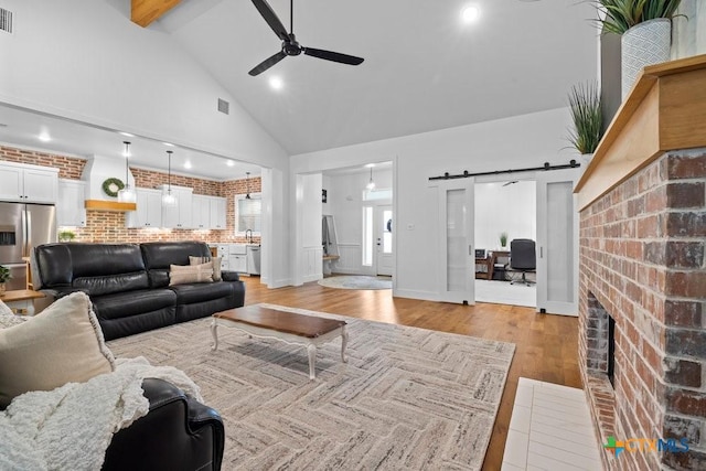 living room featuring high vaulted ceiling, light hardwood / wood-style floors, brick wall, a brick fireplace, and a barn door