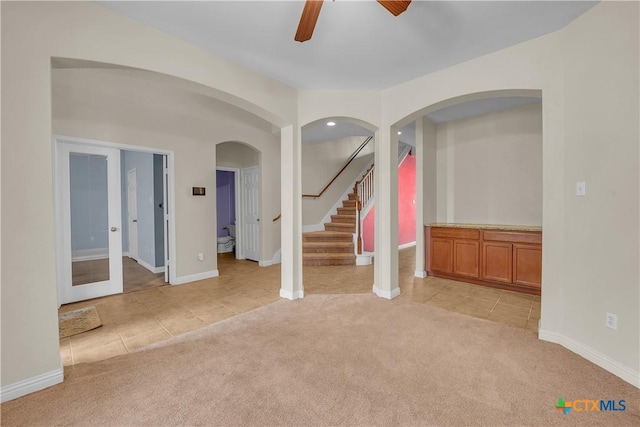 empty room featuring ceiling fan, stairs, light carpet, light tile patterned floors, and arched walkways