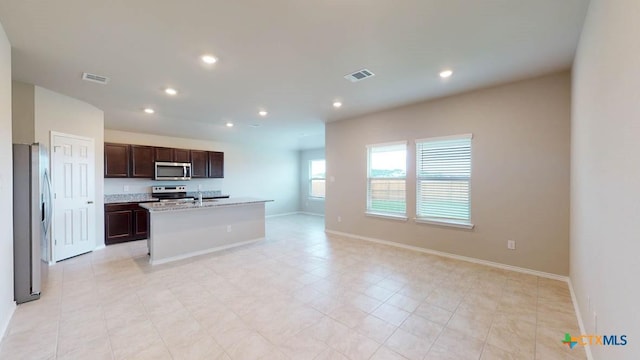 kitchen featuring an island with sink, appliances with stainless steel finishes, light stone counters, and dark brown cabinetry